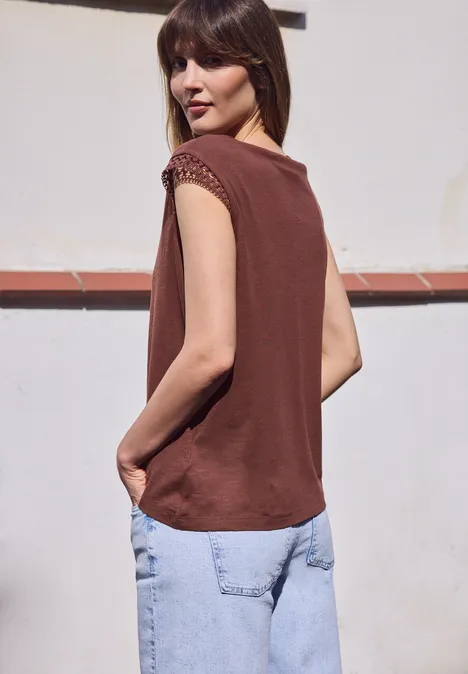 Shirt met ronde hals en gehaakte mouwen mahogany brown Vrouw in een mouwloos bruin topje en losse blauwe spijkerbroek, staand in profiel voor een witte muur.