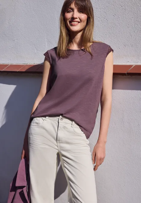 Vrouw in lila top en lichte broek voor witte muur met bijpassende tas in de hand.