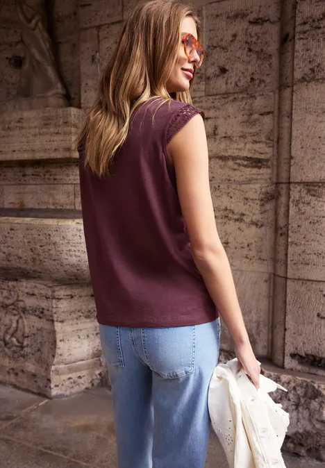 Frau von hinten in burgunderfarbenem ärmellosem Top und Jeans, hält eine helle Jacke vor einer Steinmauer.