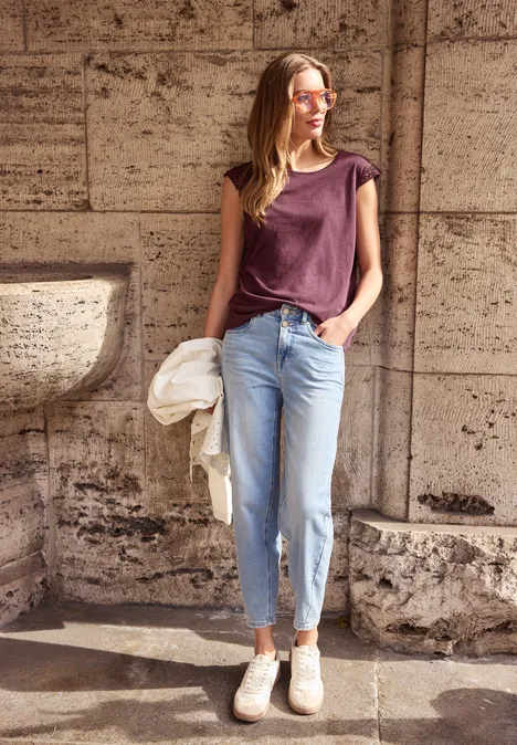 Junge Frau in Jeans und T-Shirt lehnt entspannt an einer Steinwand im Sonnenlicht.