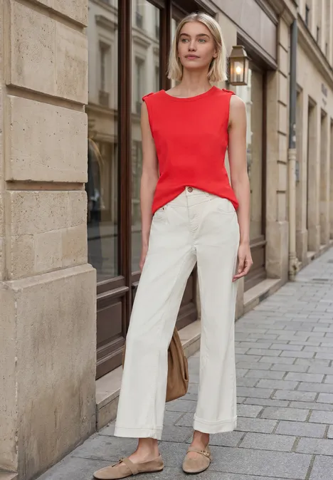 Persoon draagt rood mouwloos topje en wijde witte broek, staand op de stoep voor een historisch gebouw.
