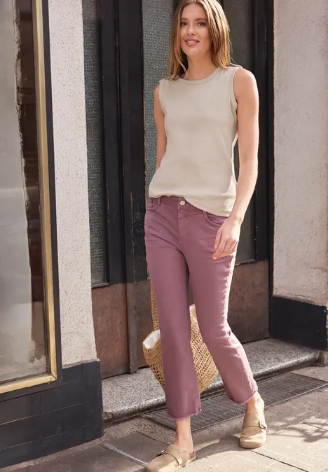 Frau in beigem Top und rosafarbenen Hosen spaziert auf sonnigem Gehweg vor Hausfassade.