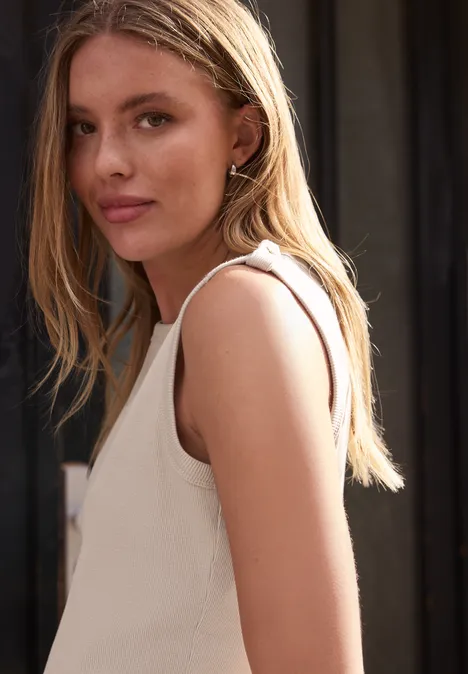 Junge Frau mit langem blondem Haar im Profil trägt ärmelloses weißes Top vor dunklem Hintergrund.