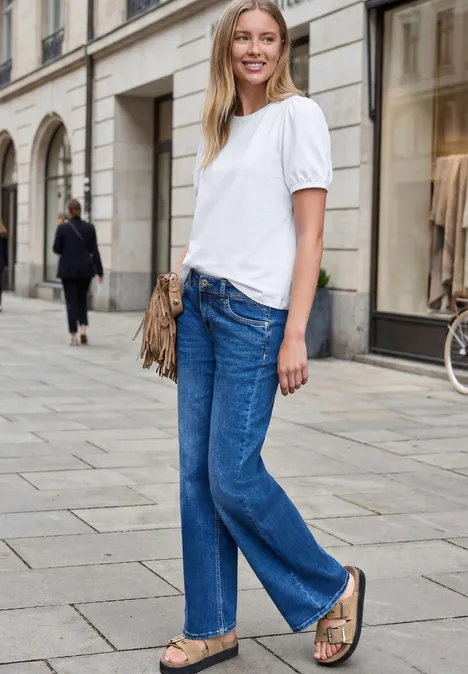Frau in weißem T-Shirt, blauen Jeans und Sandalen spaziert an einer städtischen Einkaufsstraße.