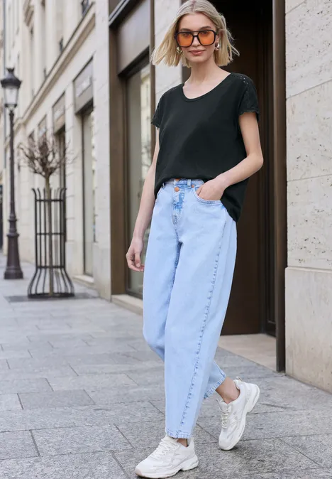 Eine Frau geht auf einer Stadtstraße, trägt schwarzes T-Shirt, hellblaue Jeans und weiße Sneaker vor einer eleganten Fassade.