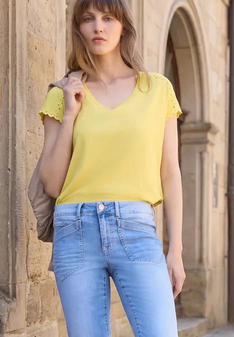 Eine Frau in gelbem Oberteil und hellblauen Jeans steht vor einem Sandsteinbogen.