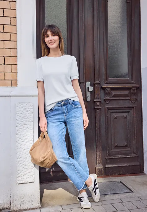 Een vrouw in een wit T-shirt en spijkerbroek leunt nonchalant tegen een muur voor een houten deur, met een tas in haar hand.