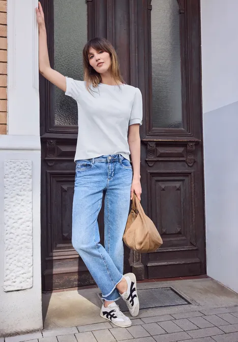 Jonge vrouw leunt tegen een deurpost, draagt jeans, wit shirt en sneakers, houdt een tas vast.
