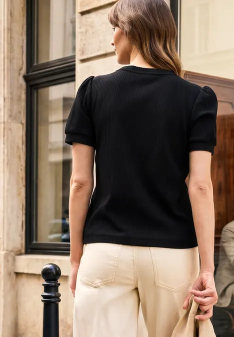 Frau steht mit dem Rücken zur Kamera, trägt schwarzen Pullover und helle Hose, vor einem Fenster.