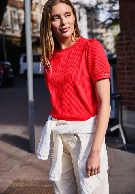 Frau in rotem T-Shirt und mit einem um die Hüfte geknoteten weißen Pullover steht auf einem Gehweg.