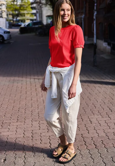 Person steht auf gepflastertem Gehweg, trägt rotes Oberteil, weiße Hose, weiße Jacke um die Hüfte und Sandalen.