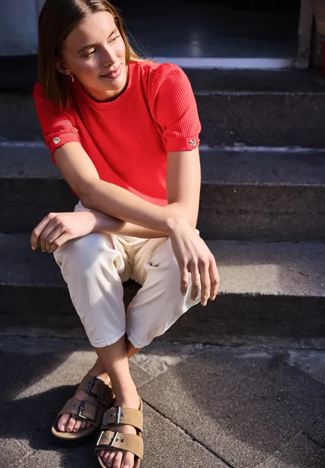 Person mit rotem T-Shirt, hellen Hose und Sandalen sitzt auf Außentreppe und verschränkt die Beine.