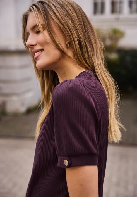 Zijaanzicht van een vrouw met bruin-achtig blond haar en een paarse trui op een stoep voor een huis.