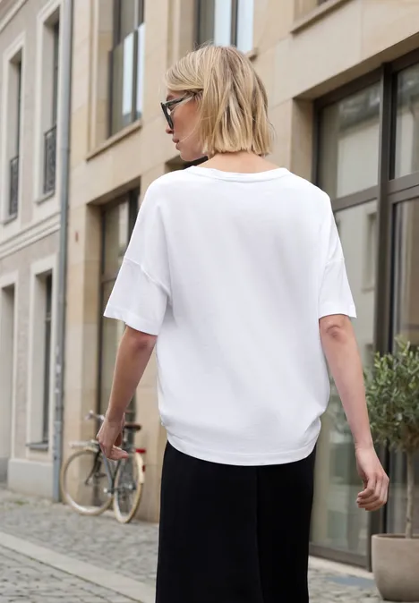 Eine Frau in weißem T-Shirt, schwarzer Rock und Sonnenbrille geht an einem Stadthaus mit Fahrrad vorbei.