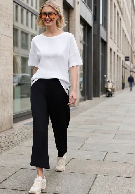 Eine Frau im weißen T-Shirt, schwarzen Hosen und weißen Sneakers läuft in der Stadt auf dem Gehweg.