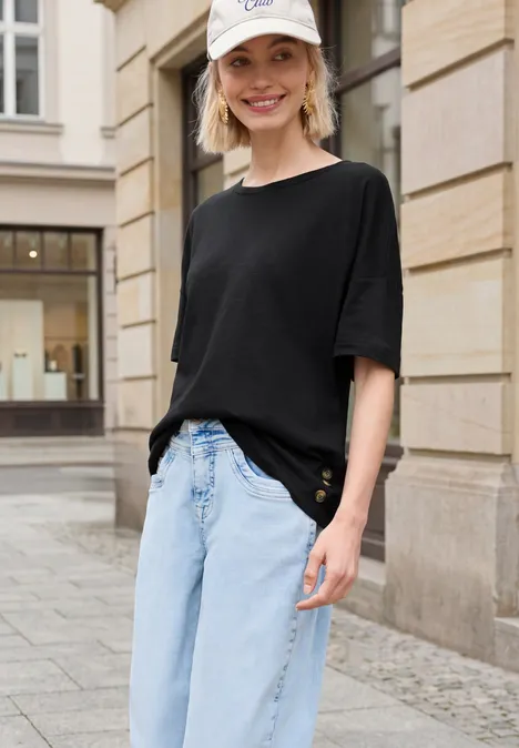 Eine Person steht vor einer Sandsteinfassade und trägt ein schwarzes Oversize-Shirt mit hellblauen Jeans.