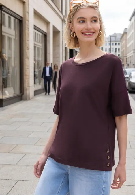 Person mit lila T-Shirt und blauen Jeans geht durch eine Einkaufsstraße, im Hintergrund Geschäfte und Passanten.