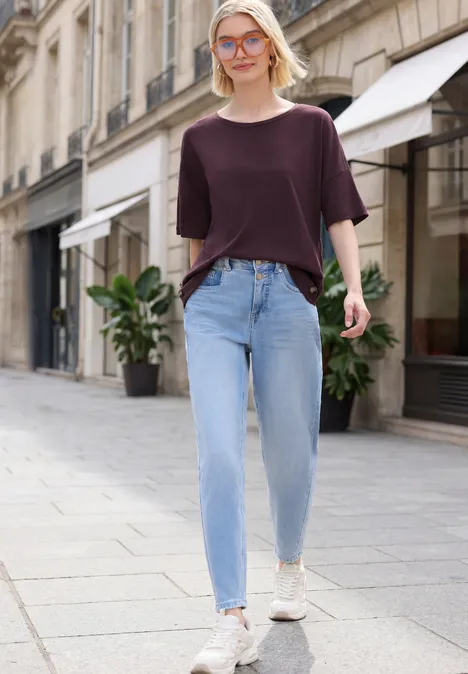 Eine Frau in einem weinroten Oberteil, hellblauen Jeans und weißen Sneakers läuft in einer städtischen Straße.