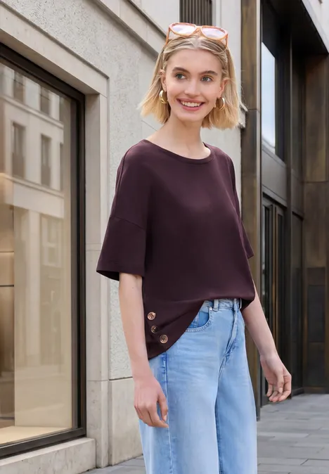 Person mit lila Oberteil und hellblauen Jeans steht vor Häuserfassade auf dem Bürgersteig.