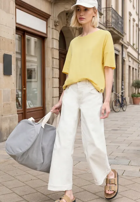 Frau in gelbem Shirt, weißen, weiten Hosen und Sandalen hält große graue Tasche auf Gehweg.