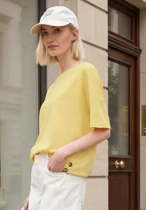 Person mit gelbem T-Shirt, weißer Hose und Cap steht vor Hausfassade.
