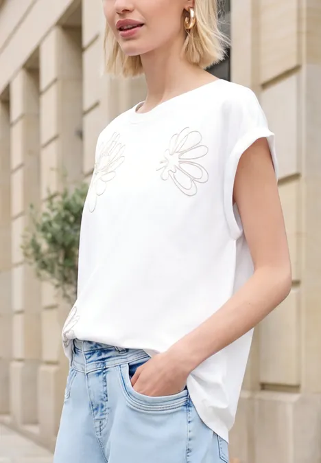 Frau in weißem, ärmellosem Shirt mit Blumenstickerei, hellen Jeans, Hand in Tasche vor Gebäude.