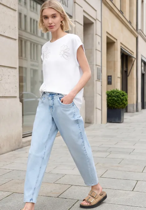 Frau in weißem T-Shirt, hellblauen Jeans und Sandalen schlendert an einer Stadtkulisse entlang.