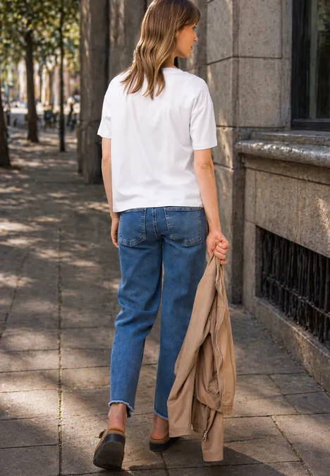 Frau in weißem T-Shirt und blauen Jeans geht mit hellbrauner Jacke in der Hand eine sonnige Straße entlang.