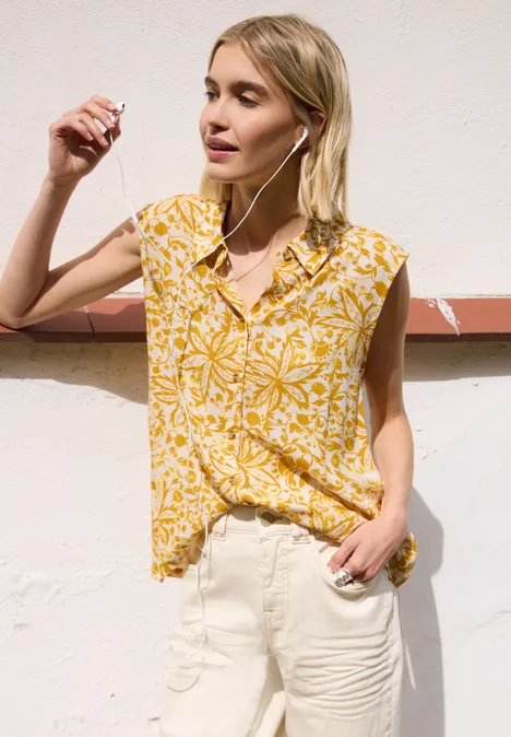 Vrouw voor lichte muur in gele bloemenblouse en witte broek.