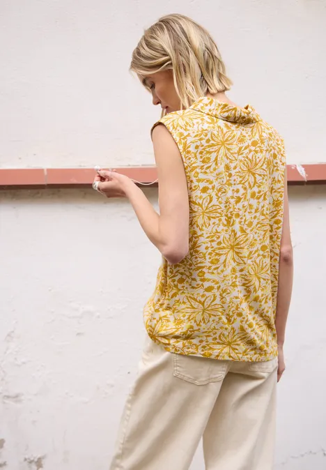 Vrouw met onscherp gezicht in een geel topje met patroon en beige broek, zijaanzicht voor een witte muur.