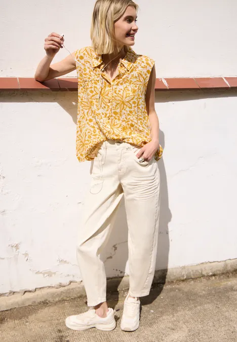 Persoon leunt tegen een witte muur, draagt een gele top met print, lichte broek en witte sneakers.