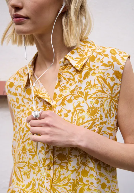 Vrouw met blond haar draagt een mouwloze, geel-witte bloementop en oordopjes