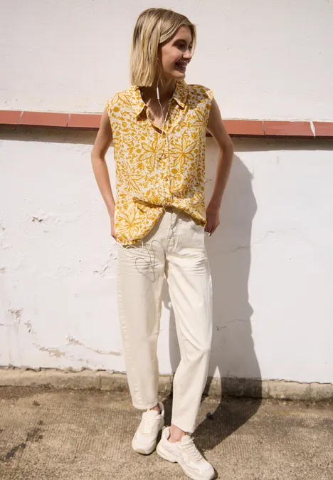 Persoon leunt tegen lichte muur, draagt gele gebloesde top, lichte broek en witte sneakers.