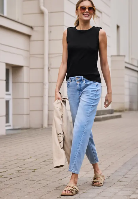 Vrouw in zwarte top, blauwe jeans en sandalen, met een jas in de hand, lopend op de stoep.