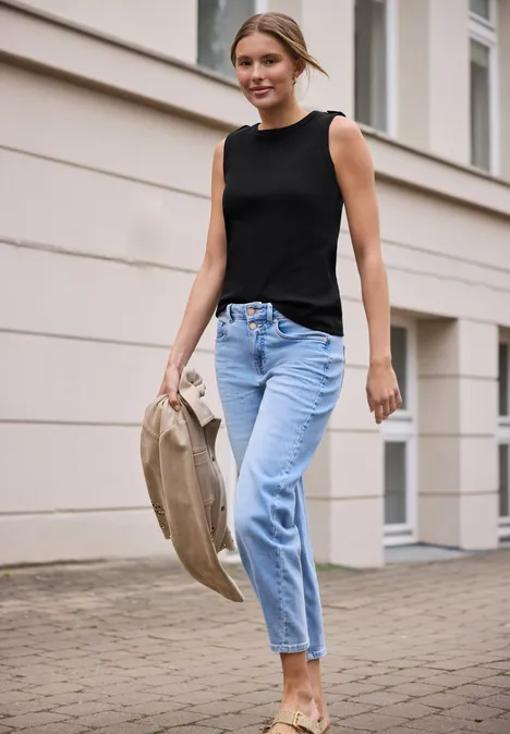 Vrouw in zwart topje en lichtblauwe jeans loopt langs een gebouw met een beige hoed in haar hand.