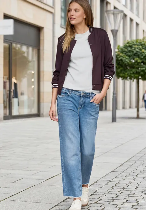 Eine Frau in Jeans, weißem Shirt, Bomberjacke und Sneakern geht an Geschäften in der Stadt vorbei.