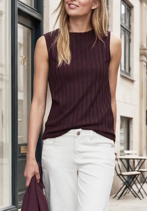 Frau in ärmellosem, bordeauxfarbenem Top und weißen Jeans hält einen Pullover in der Hand.
