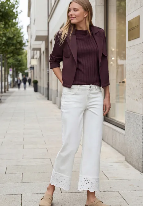 Eine Frau mit verschwommenem Gesicht steht auf dem Gehweg in weißer Spitzenhose und lila Oberteil.