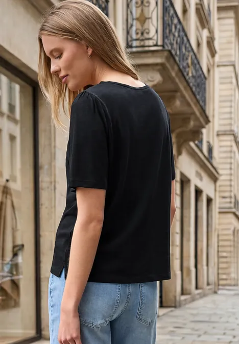Eine Frau in schwarzem T-Shirt und blauen Jeans steht seitlich auf einer gepflasterten Straße vor historischen Gebäuden.