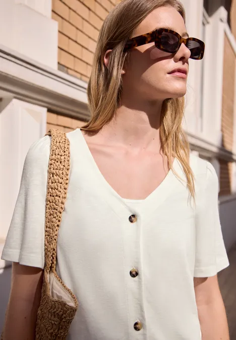 Frau in weißem kurzärmligem Stricktop mit Knöpfen und Basttasche vor Hausfassade