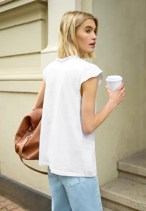 Persoon van achteren met wit T-shirt, jeans en koffiebeker, met een bruine tas voor een gevel.