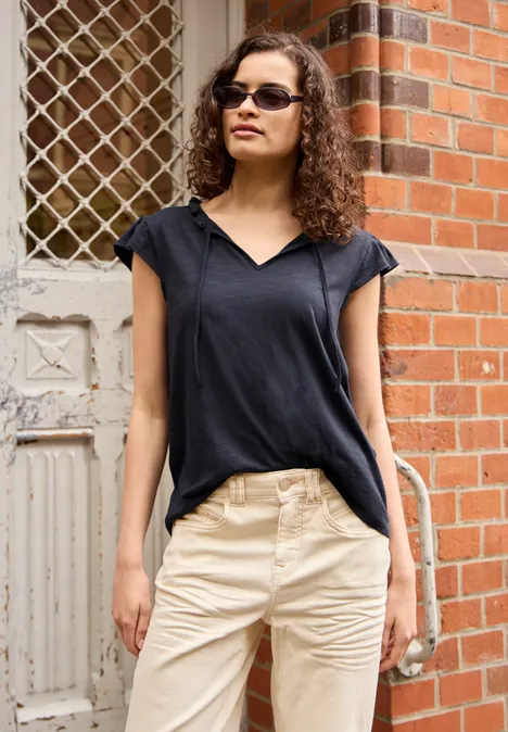 Vrouw in zwart shirt en beige broek voor een bakstenen muur.