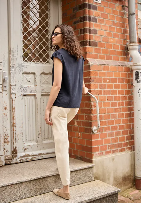 Vrouw met krullend haar en zonnebril, donkerblauw shirt, lichte broek en beige tas beklimt een trap.