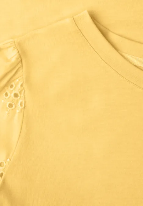 Nahaufnahme eines gelben T-Shirts mit Rundhalsausschnitt und filigranen Lochstickereien an der Schulter.
