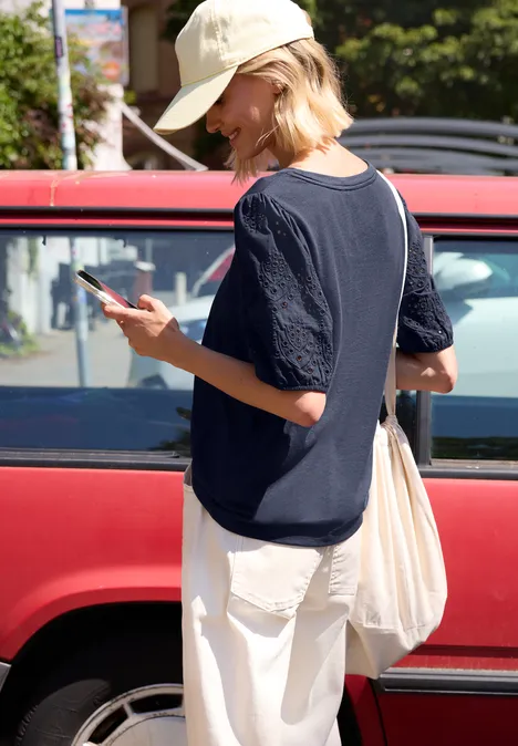 Frau in Kapuzenpulli, weißer Hose und Mütze blickt auf ihr Handy neben rotem Auto.