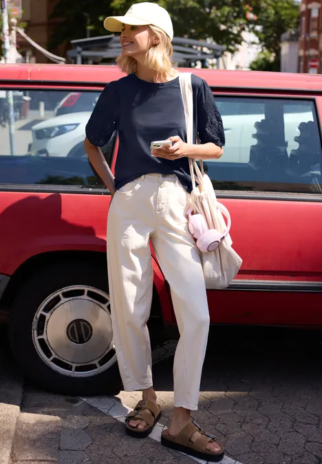 Person in T-shirt, weiten Hosen und Sandalen steht vor rotem Auto, hält Smartphone und hat Tasche mit Kuscheltier.