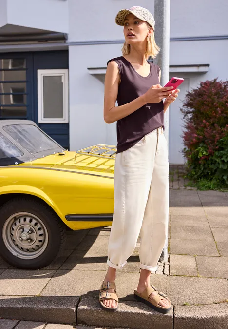 Vrouw in losse kleding houdt een smartphone vast naast een gele sportauto in woonwijk.