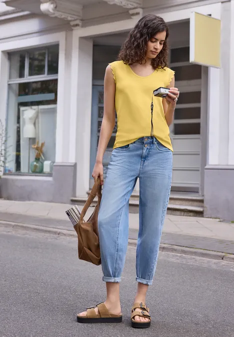 Top mit V-Neck und Rüschen snap yellow Frau in gelbem T-Shirt, blauen Jeans und Sandalen, hält Smartphone und braune Tasche auf der Straße.