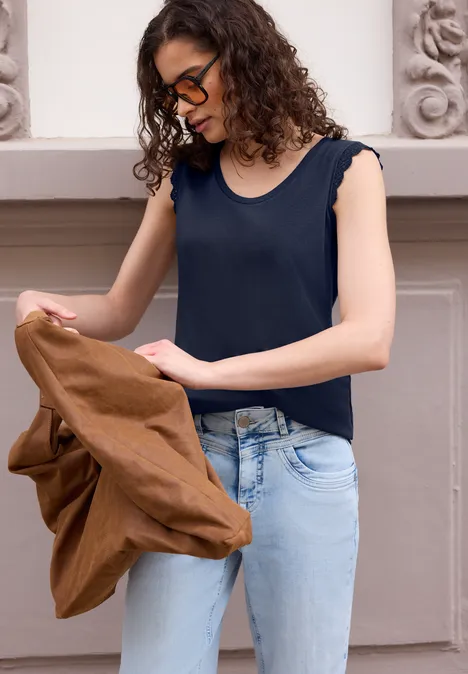 Frau mit lockigem Haar in schwarzem Top und Jeans hält eine braune Jacke vor einer Hauswand.