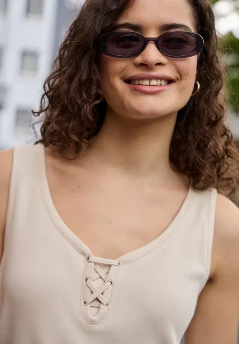 Geschlechtsneutrale Person mit lockigem Haar trägt hellbeiges, ärmelloses Top mit Schnürung, Gesicht unscharf.